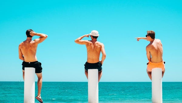 Men wearing swimwear, sitting at the beach looking out to sea. 